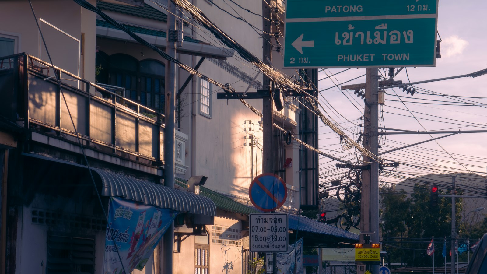 A street in Phuket with a green road sign reading เข้าเมือง, Phuket Town, above shophouses and tangled power lines.