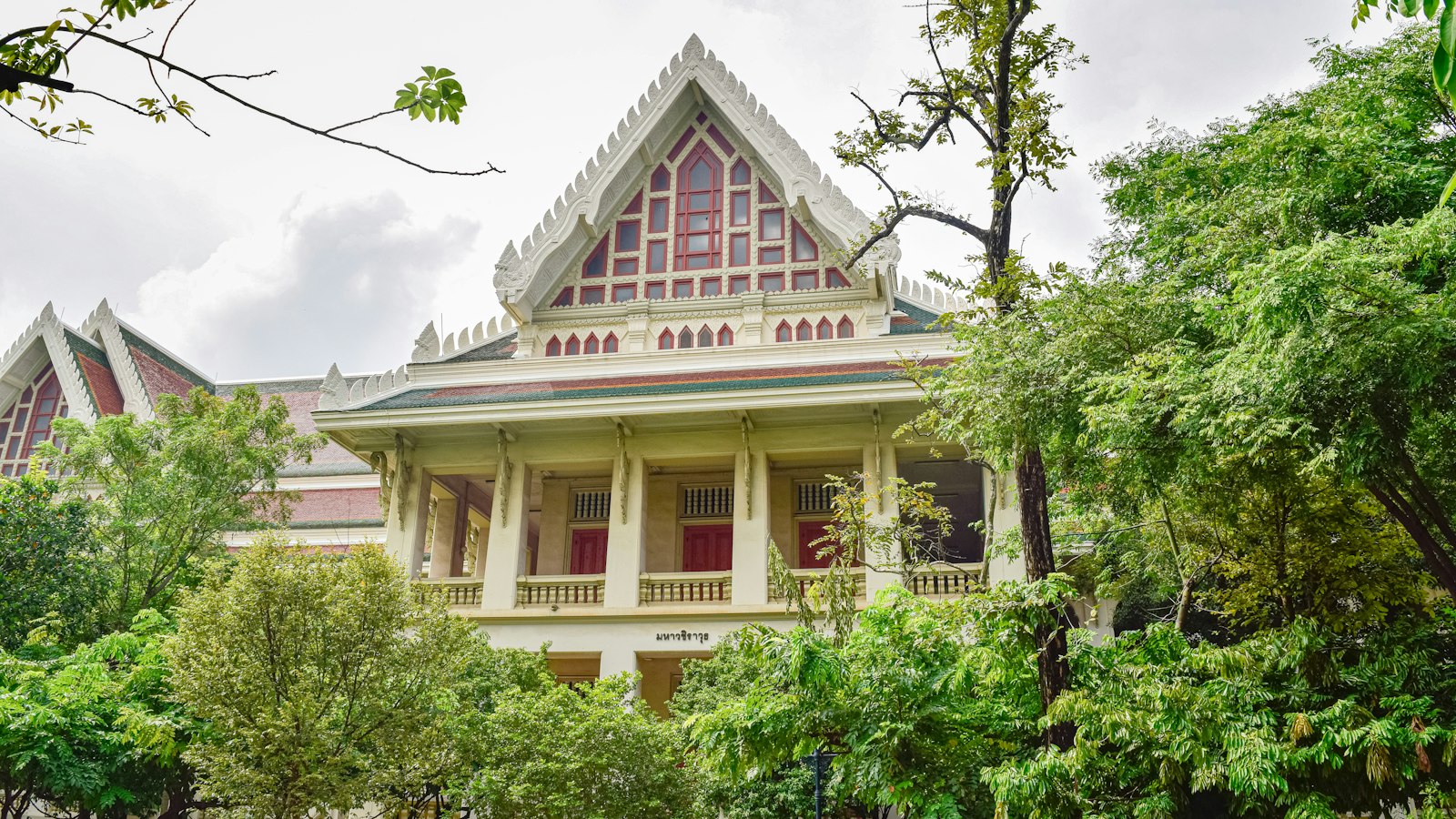 A white university building with a tiered red-and-white Thai-style roof, half hidden by trees, in Bangkok.