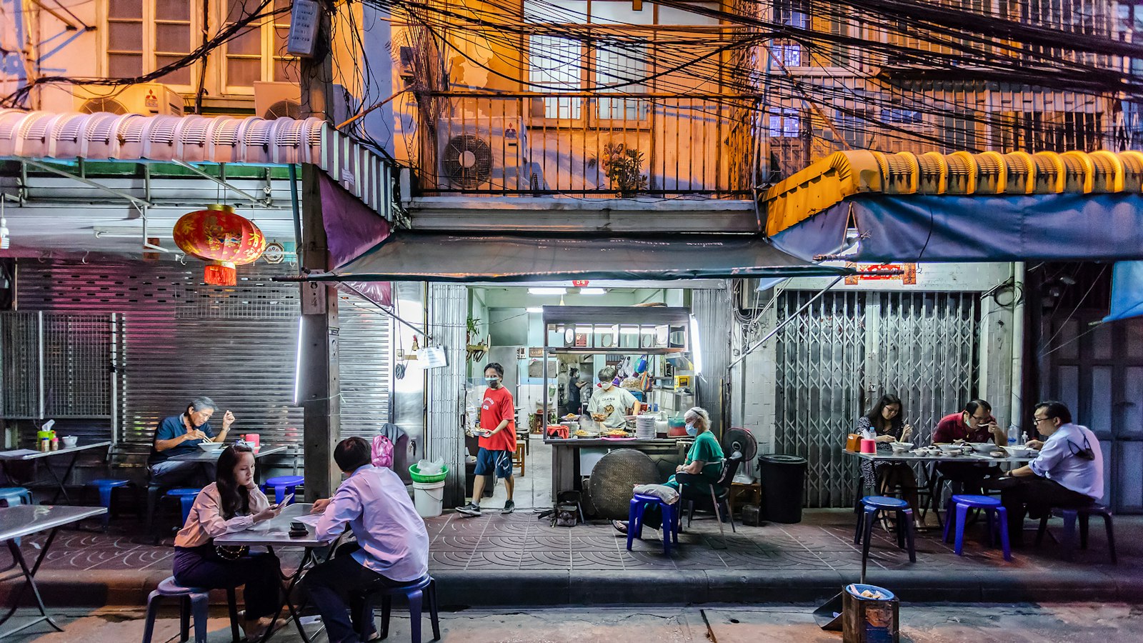 People eating and chatting at plastic tables outside a street food shop in Bangkok at night.